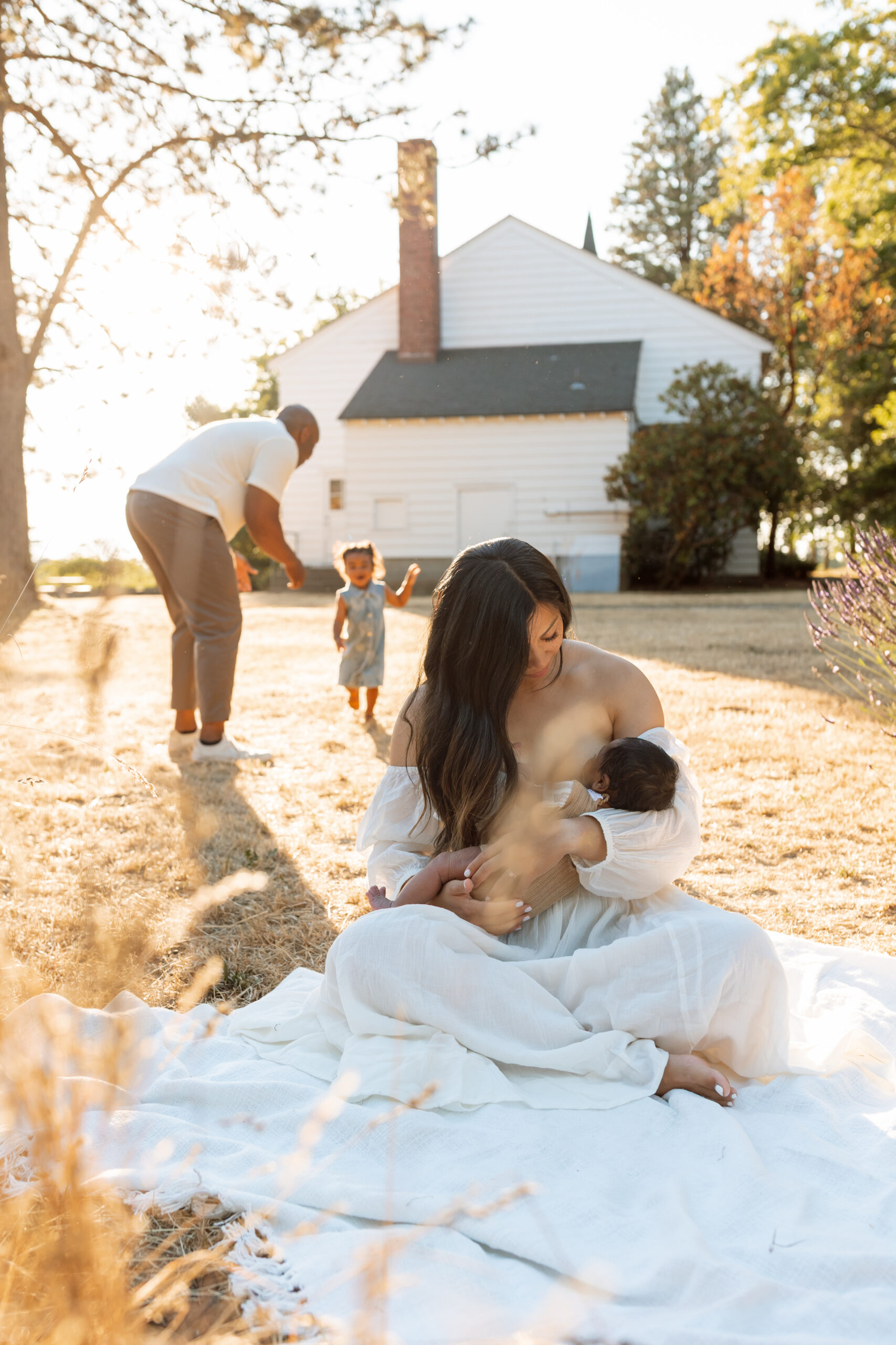 Mother cuddling her newborn outdoors while a toddler and father play nearby during a family session