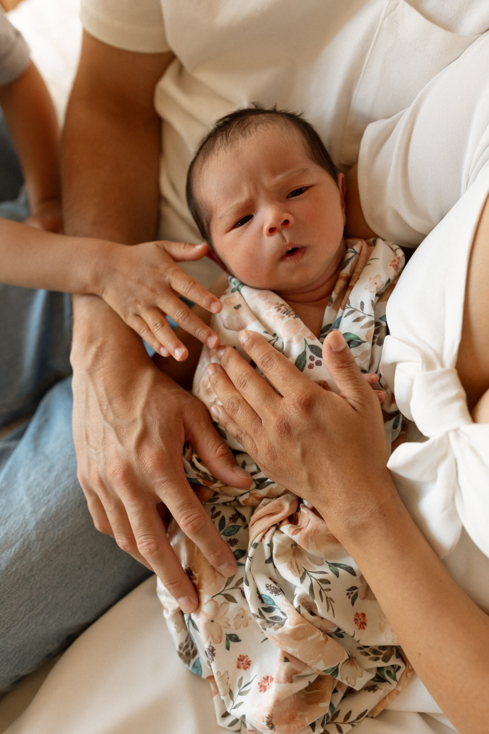 Close-up newborn photography in Seattle featuring a baby wrapped in a floral swaddle surrounded by parents' and sibling's hands.